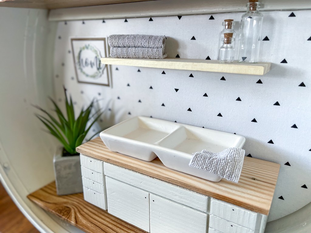 Kitchen in a dollhouse made in a vintage suitcase complete with double sink, floating shelf, and tiny towels.