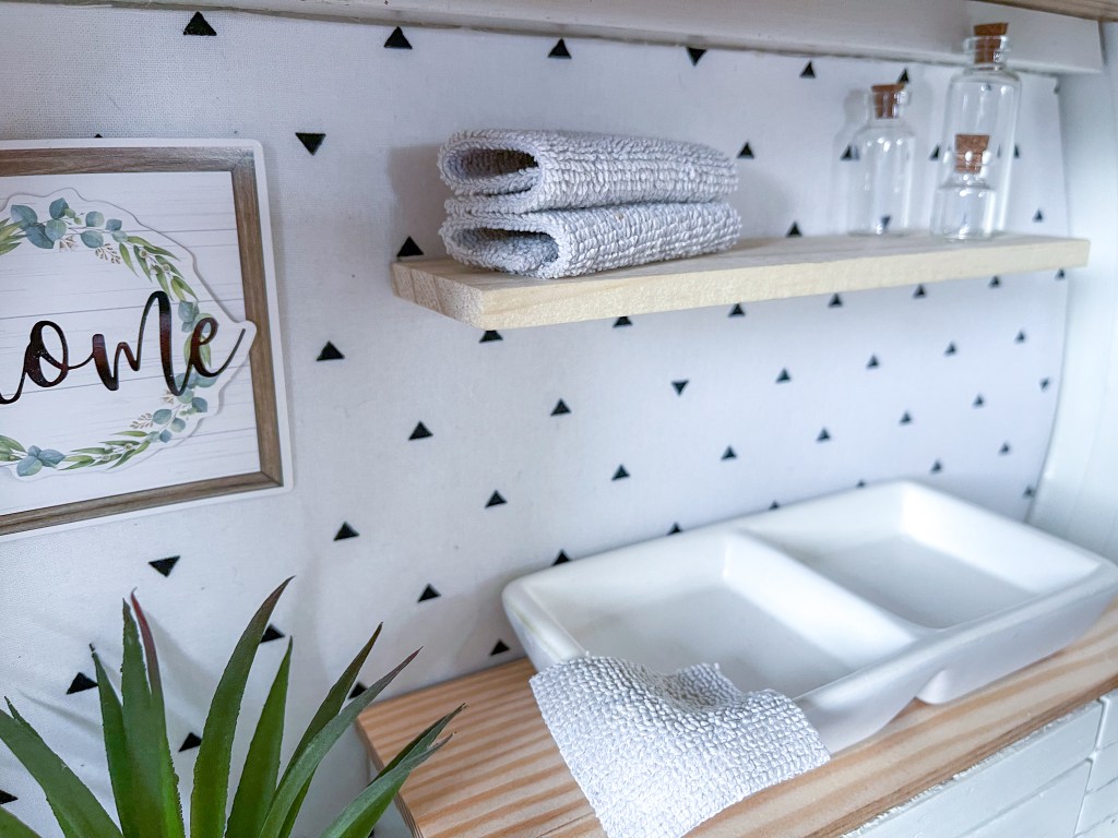 Close-up of tiny kitchen with double sink, floating shelves, glass jars and towels in a dollhouse made in a vintage suitcase.