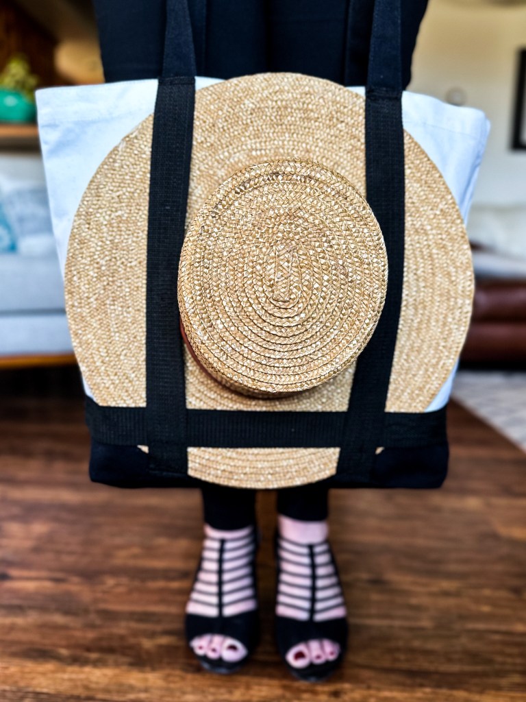A woman strolls along, her black strappy shoes clicking softly, as she carries a stylish sun hat tote bag, showcasing the perfect blend of functionality and summer fashion